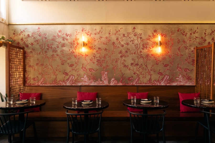 Elegant dining area with a floral mural on the wall, featuring two round tables set for service and cushioned seating against the wall with warm ambient lighting.