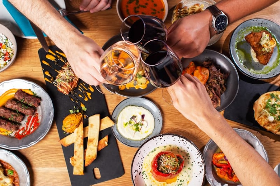 Hands holding glasses of wine over a table filled with various dishes, including appetizers, dips, and entrees.