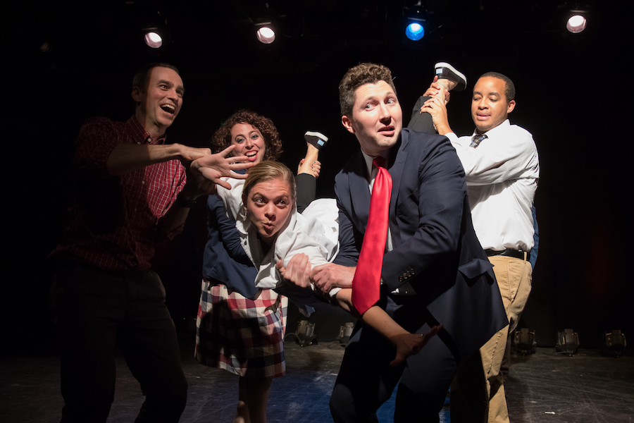 Actors perform a lively scene on stage, with one actor being held up by others. The group is dressed in casual and formal attire, performing energetically under stage lights.