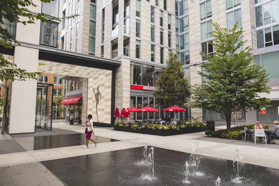 A person walking through a courtyard at CityCenterDC, with water fountains in the foreground and outdoor seating with red umbrellas near a restaurant in the background.