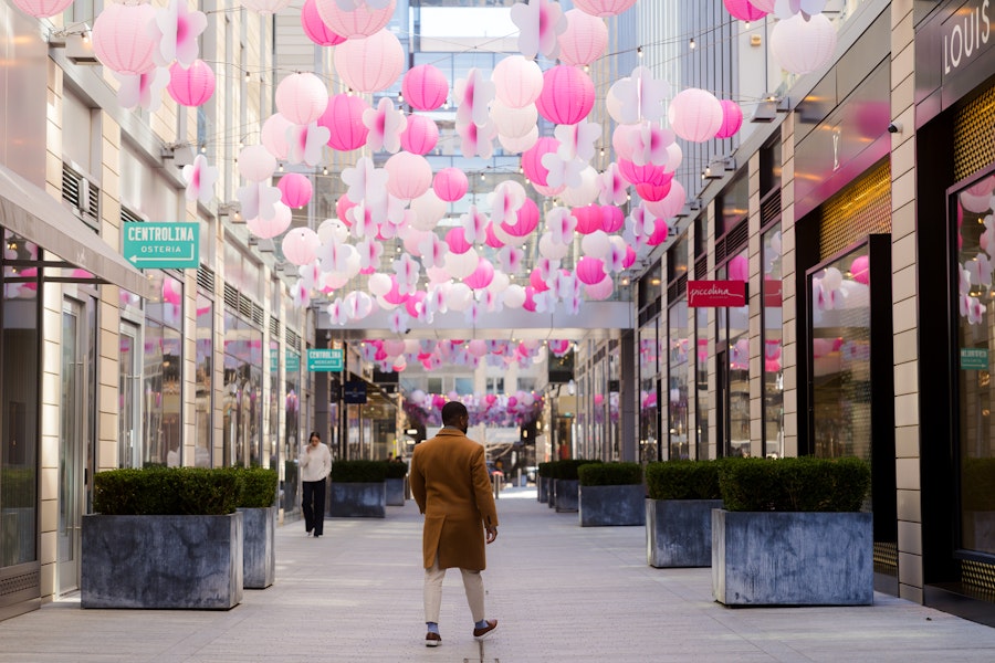 A person in a brown coat walking through a CityCenterDC passageway adorned with pink and white lanterns hanging above, with storefronts on either side.