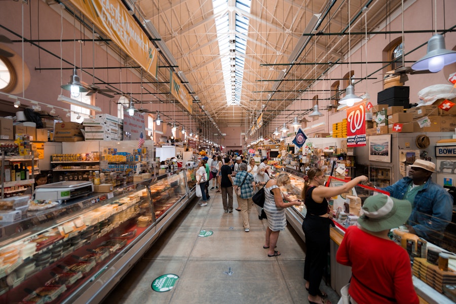 The interior of Eastern Market is lively with vendors and shoppers. The long aisle is lined with stalls offering an array of products, and the high ceiling is supported by a framework of beams.