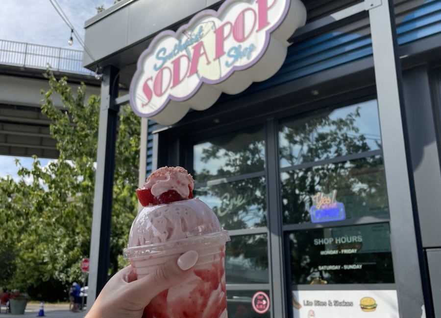 A hand holding a cup of strawberry ice cream in front of the “Southwest Soda Pop Shop” sign.