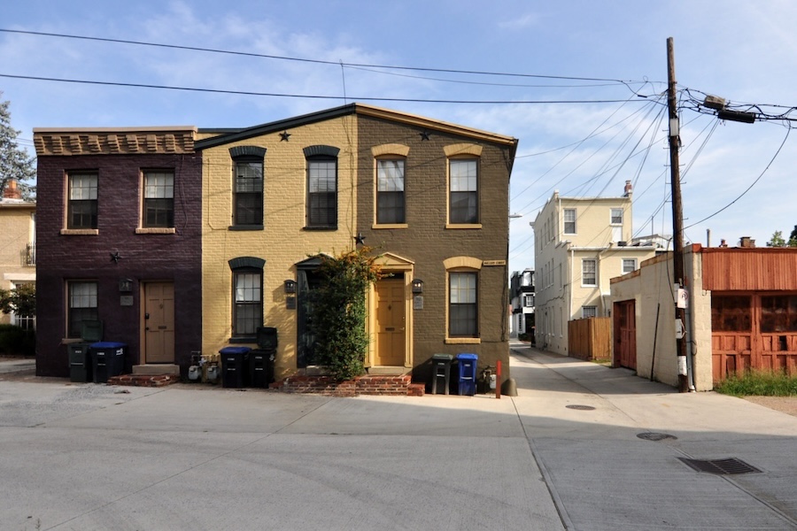 A pair of brick townhouses on a quiet street corner, with one painted yellow and the other painted dark red, connected by a shared wall. Utility poles and wires are visible above, and trash bins are lined up in front of the homes.