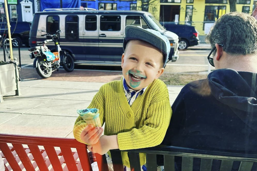  A smiling child with blue ice cream smeared on their face, holding an ice cream cone, sitting on a bench.