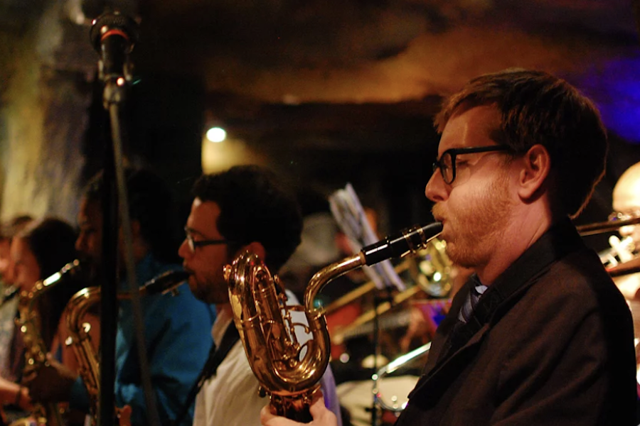 A group of musicians playing brass instruments, including saxophones, perform in a dimly lit venue. The musicians are focused on their music, with one man in glasses in the foreground playing a saxophone.