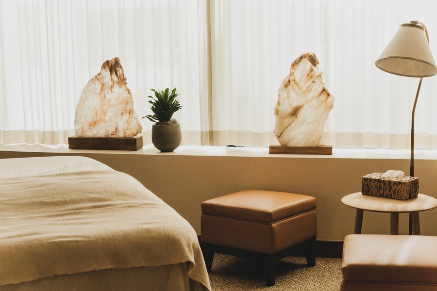 A serene therapy room with soft lighting features a neatly made bed, two large crystal sculptures on the windowsill, and warm, minimalistic decor including leather stools and a small table.