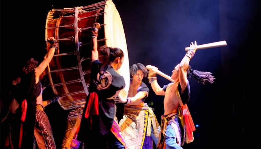 A group of dynamic performers from Yamato, a Japanese taiko drumming ensemble, energetically play a large drum, with one drummer striking it while others hold it in place, all dressed in traditional garments and illuminated by dramatic stage lighting.