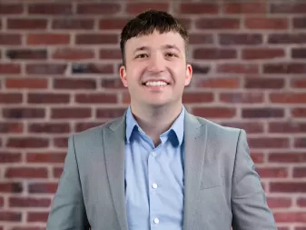 A man with short dark hair, wearing a light gray blazer over a blue button-up shirt, stands in front of a brick wall.